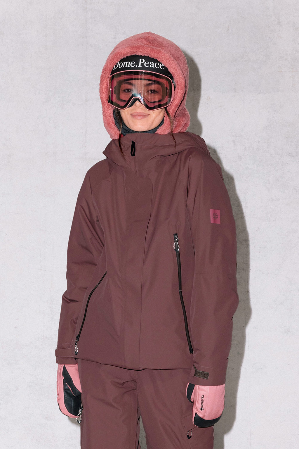 A person stands against a light gray background, smiling slightly in the 686 Women's Whisper™ Insulated Jacket and pants, pink gloves, a fuzzy pink hood, and ski goggles with "Dome. Peace" on the strap.