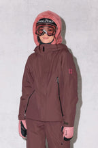 A person stands against a light gray background, smiling slightly in the 686 Women's Whisper™ Insulated Jacket and pants, pink gloves, a fuzzy pink hood, and ski goggles with "Dome. Peace" on the strap.