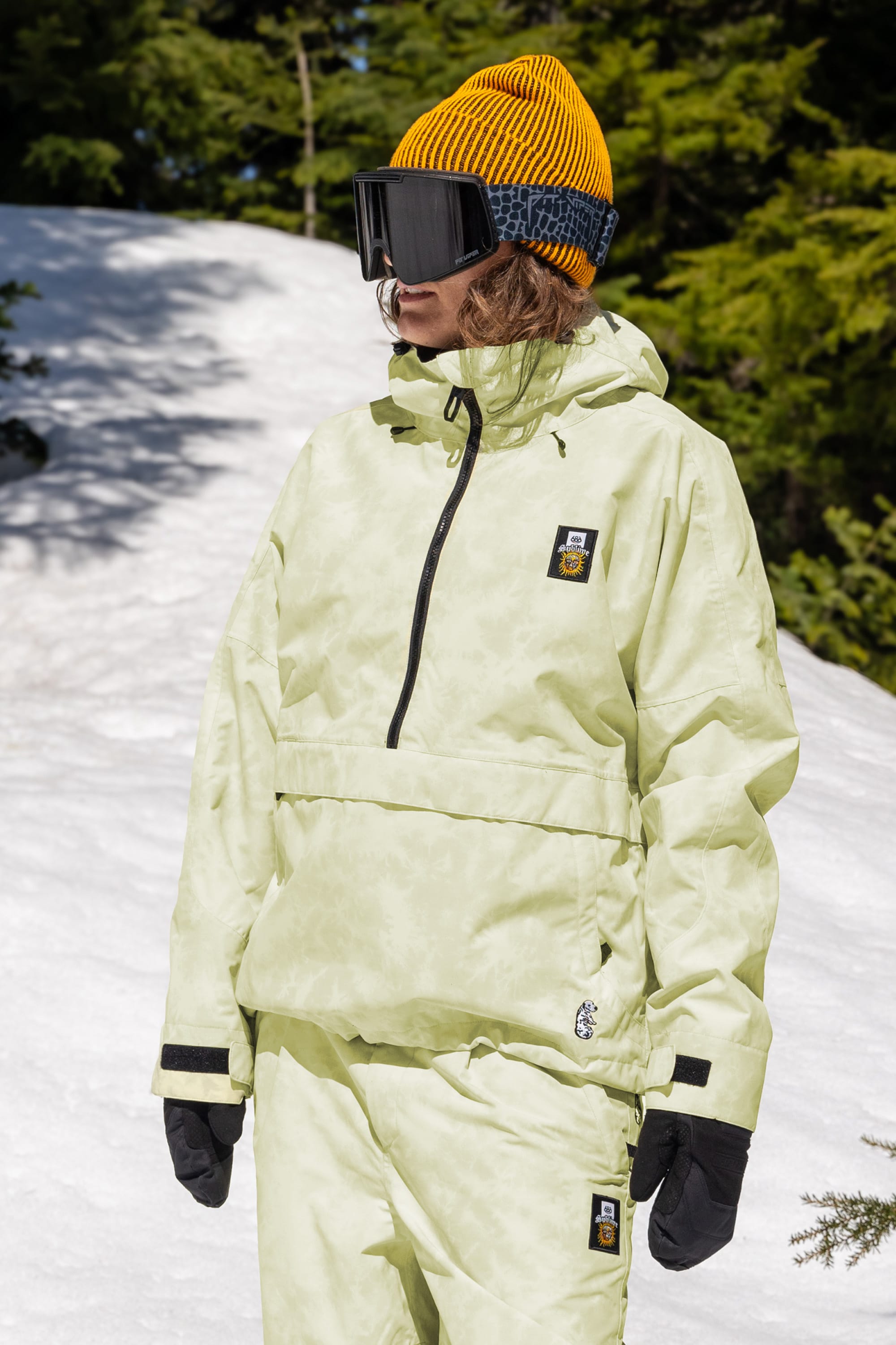 Wearing the 686 Womens Sublime Shell Anorak, a person in black gloves, yellow beanie, and ski goggles stands on snow with green trees behind them.