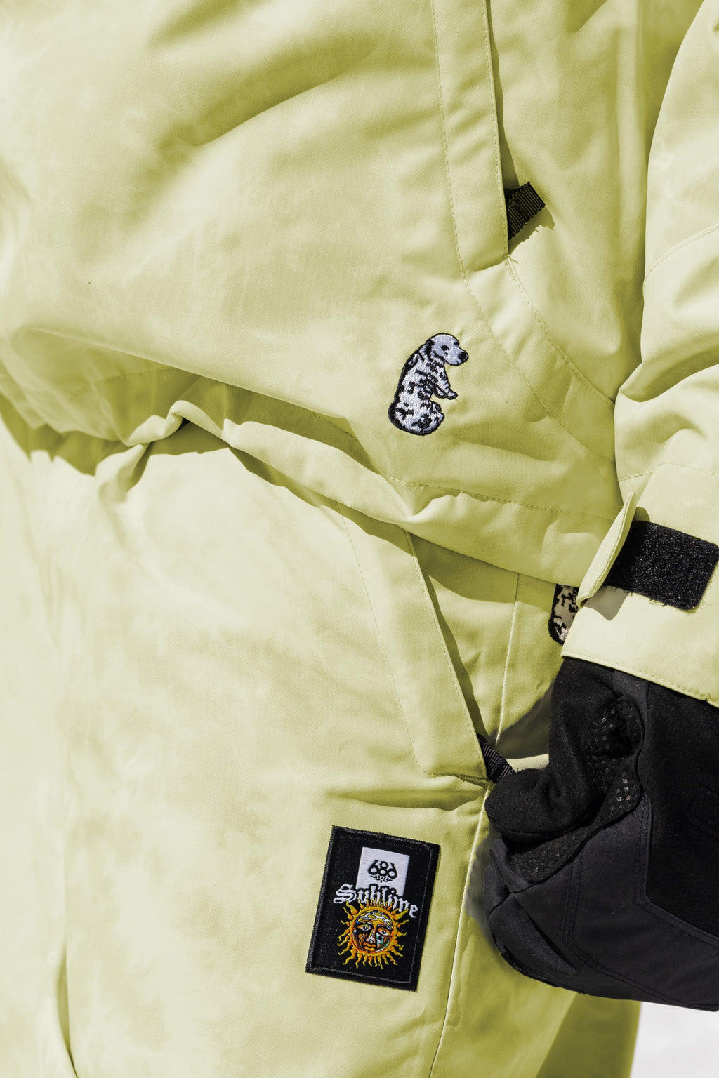 A person wears the 686 Womens Sublime Shell Anorak in pale yellow with matching pants, featuring visible patches—a small black and white dog, a sun with “Sublime”—and accessorizes with a black glove.