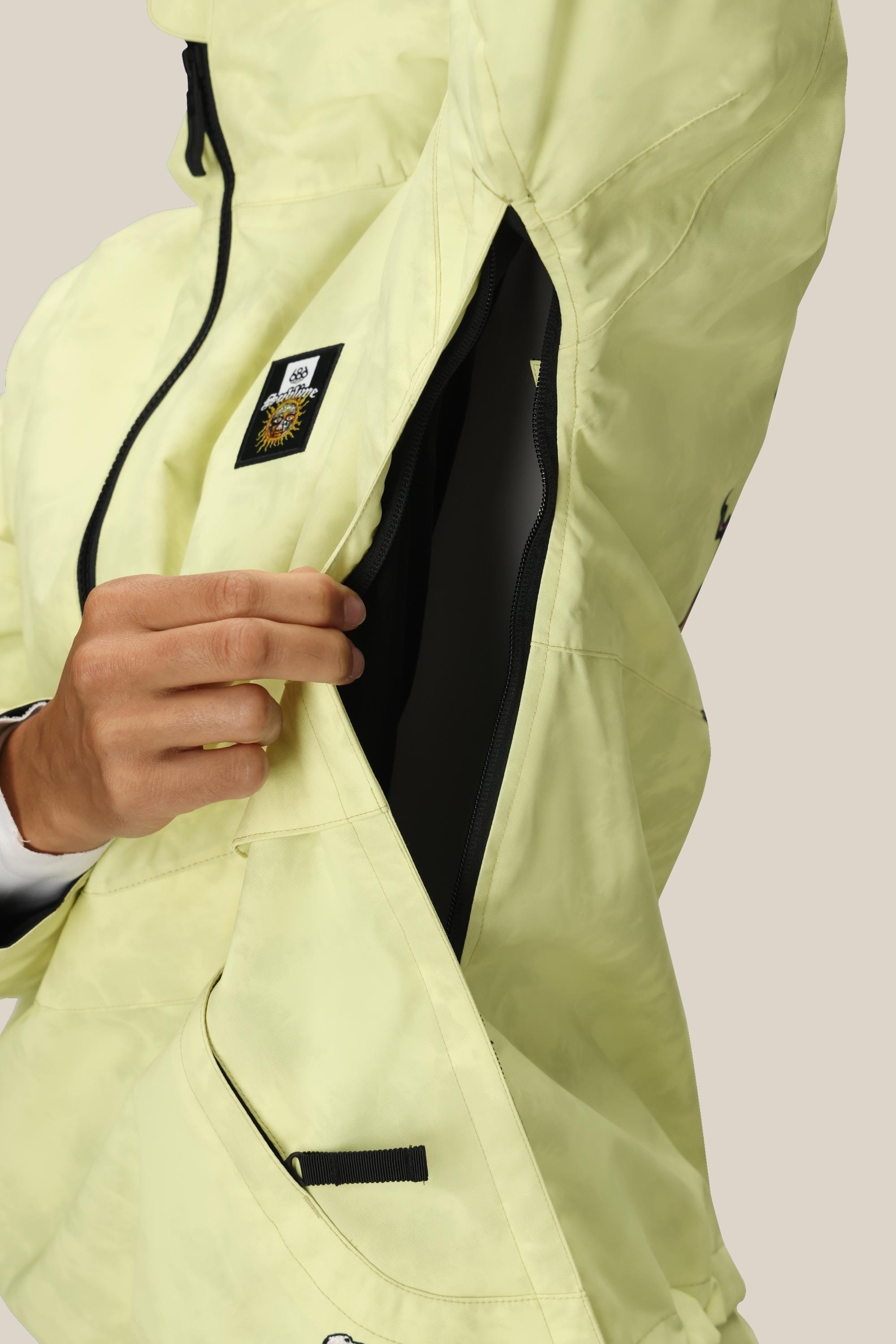 A person wearing the 686 Womens Sublime Shell Anorak in light yellow unzips a large side vent, revealing the black interior lining. The jacket features a black and yellow patch near the chest.