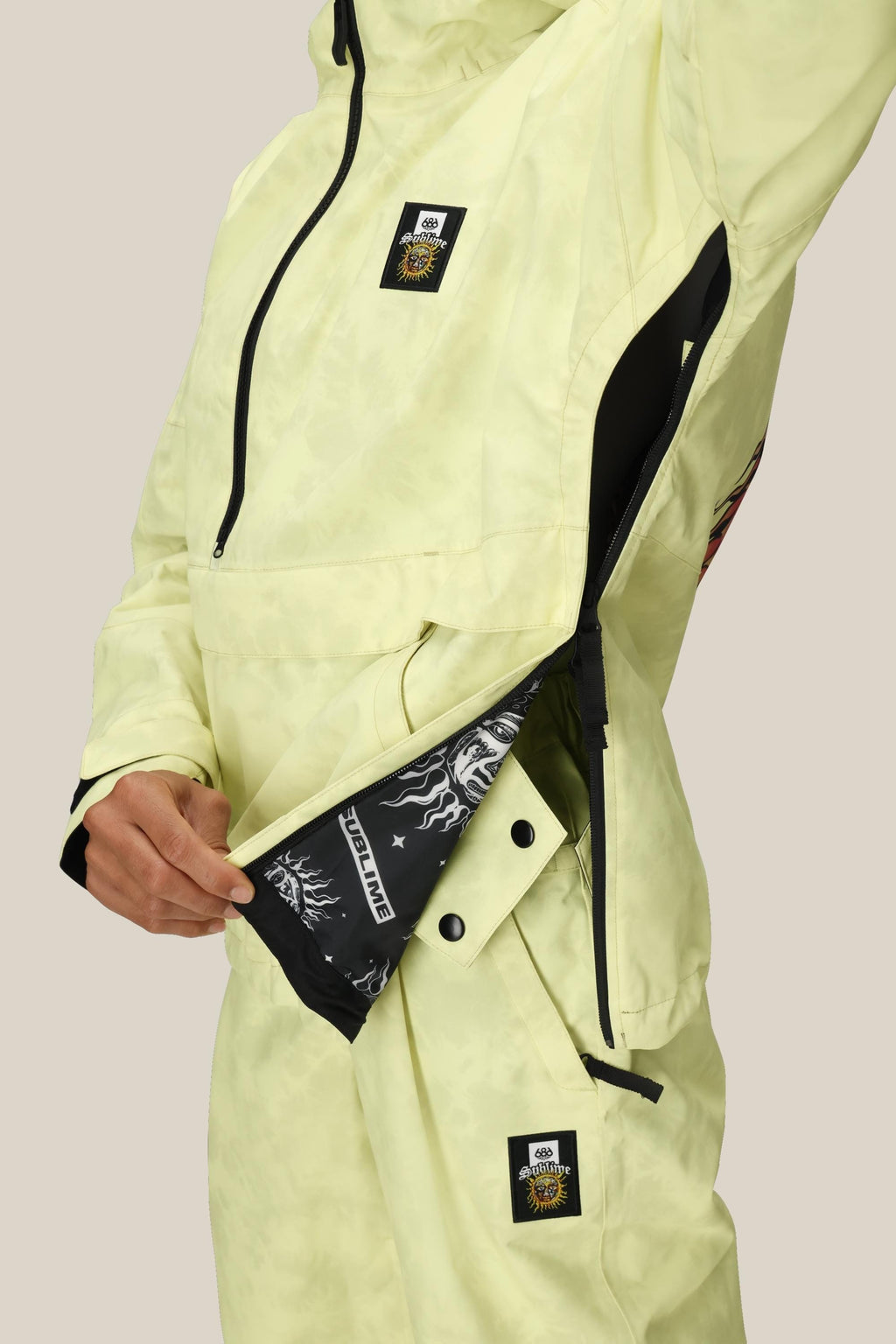 A person wearing the 686 Womens Sublime Shell Anorak unzips a large side zipper, revealing a black inner lining with white graphic designs. The pale yellow jacket and pants feature square black-and-white patches on the chest and pants.