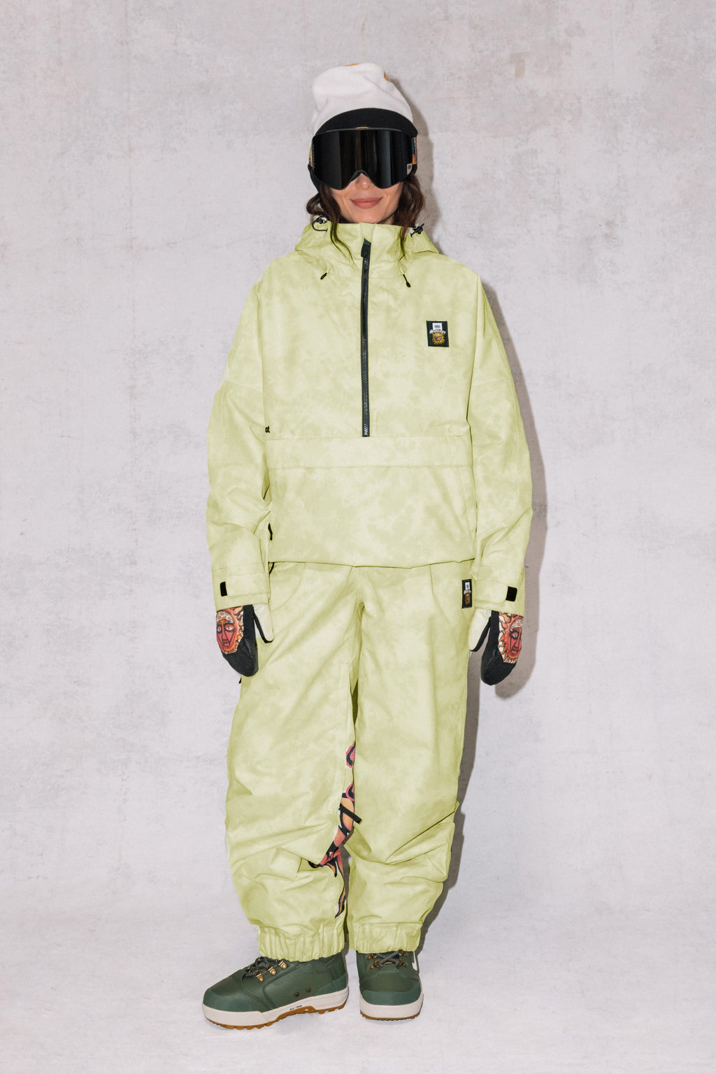 A person wears the 686 Womens Sublime Shell Anorak in light yellow-green with matching pants, dark gloves, green boots, a white beanie, and black ski goggles, standing against a light gray textured background.