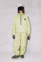 A person wears the 686 Womens Sublime Shell Anorak in light yellow-green with matching pants, dark gloves, green boots, a white beanie, and black ski goggles, standing against a light gray textured background.