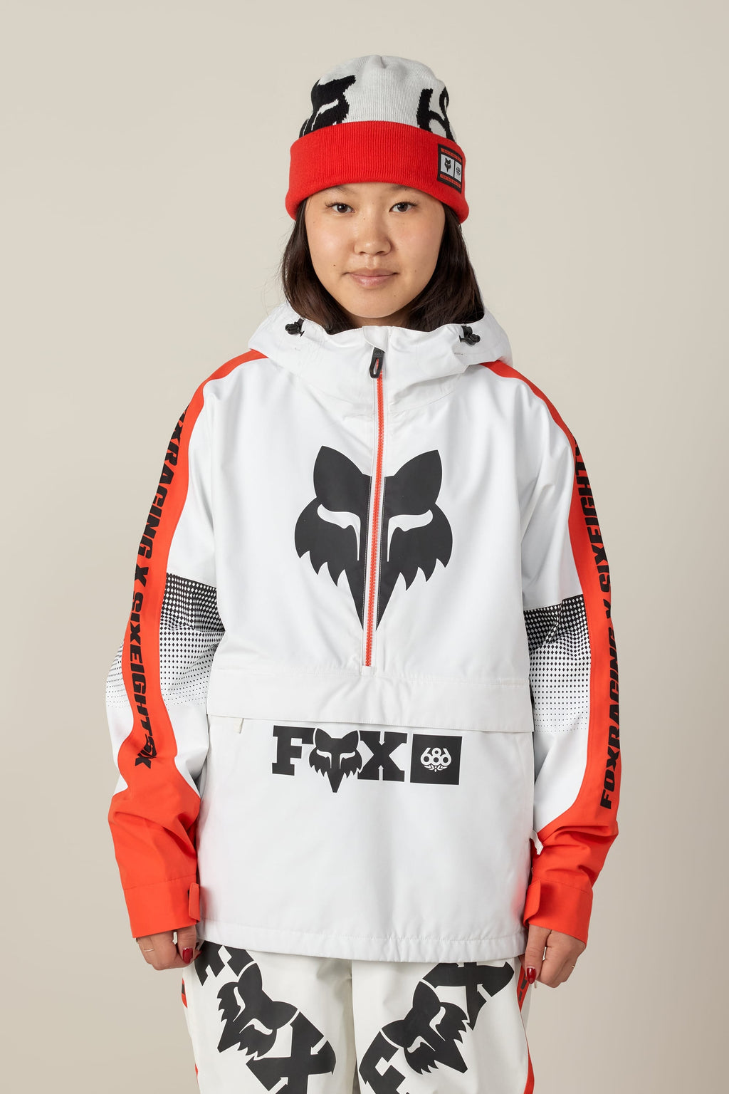 A person wearing the 686 Women's Fox Racing Shell Anorak, featuring a bold black fox logo, poses against a plain beige background.