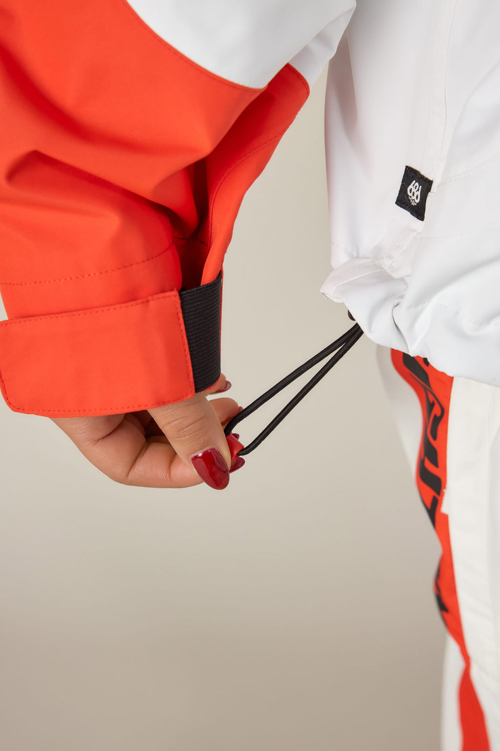 A person with red nails adjusts the drawstring of the 686 Women's Fox Racing Shell Anorak. The close-up highlights their hand, sleeve, and the details of the white and orange jacket from 686.