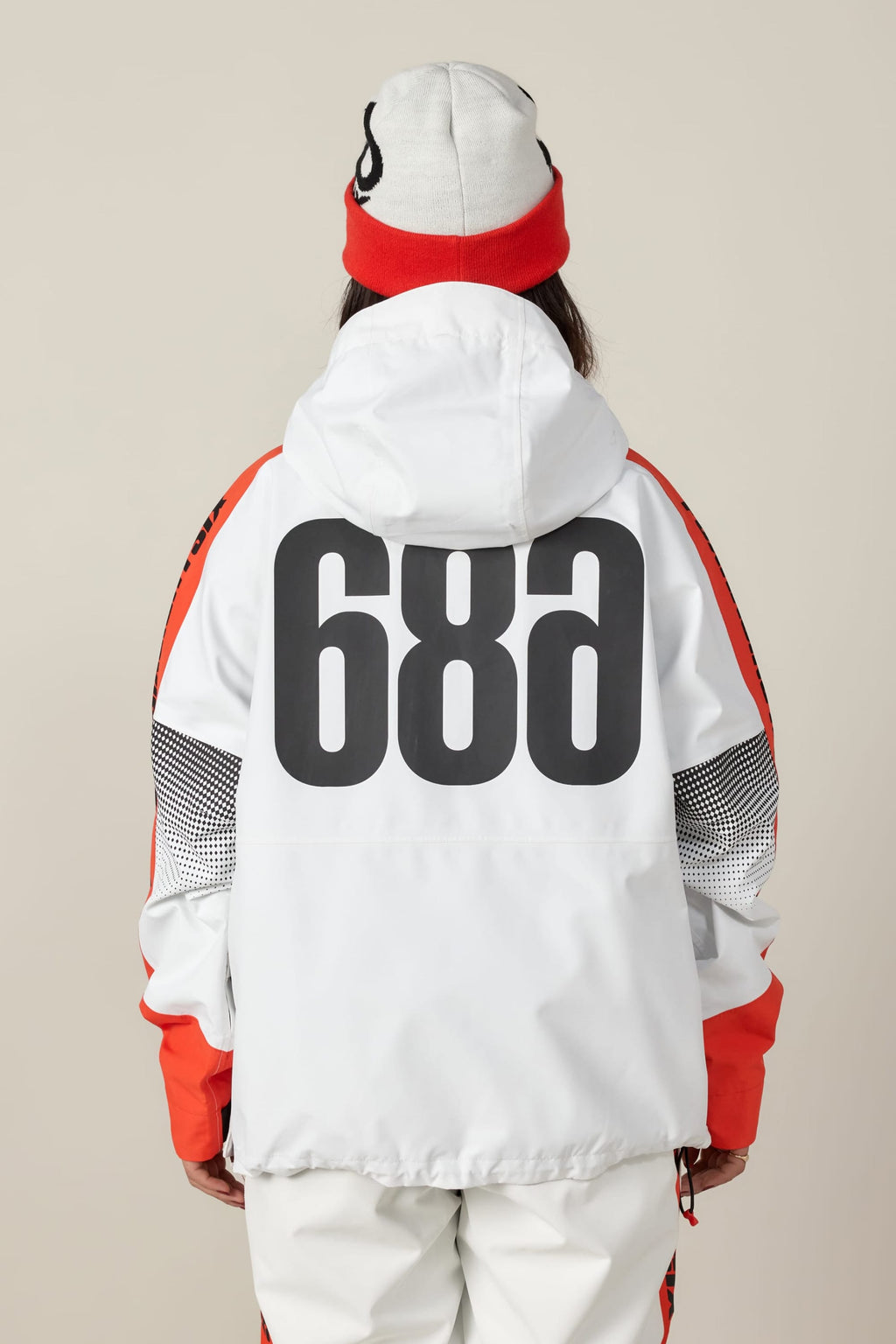 A person wearing the 686 Women's Fox Racing Shell Anorak by 686, featuring white and red colors and bold black "68a" lettering on the back, with white pants and a matching beanie, facing away against a beige background.