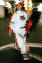 Wearing the 686 Women's Fox Racing Shell Anorak by 686, a person walks indoors holding a snowboard. The motion blur effect adds dynamic energy to the scene.