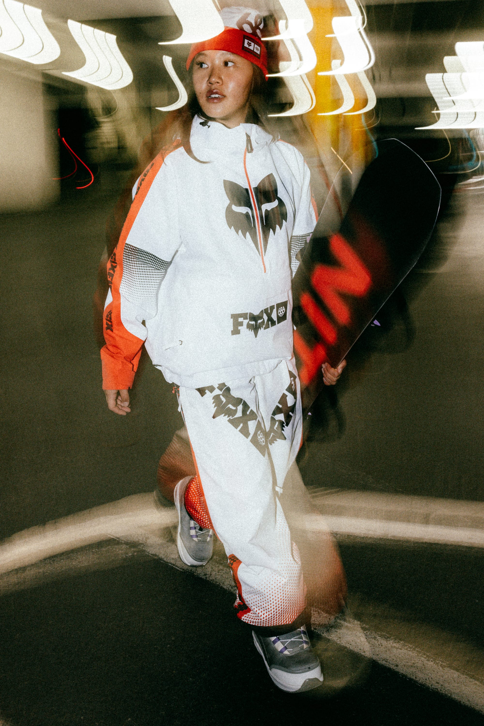 Wearing the 686 Women's Fox Racing Shell Anorak by 686, a person walks indoors holding a snowboard. The motion blur effect adds dynamic energy to the scene.