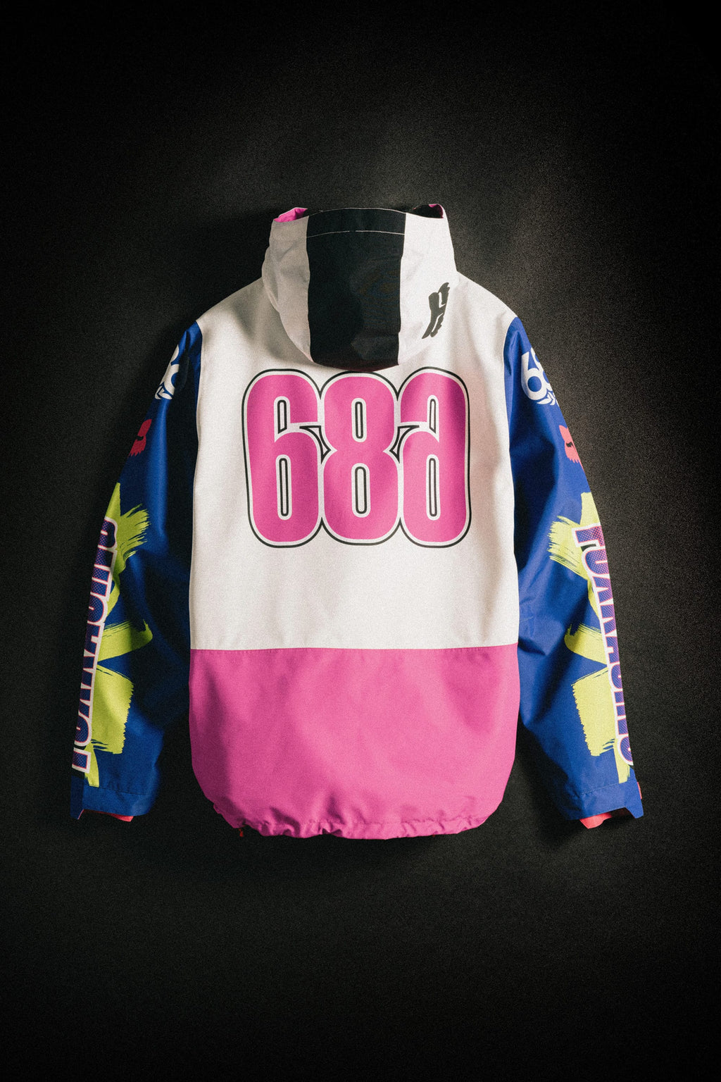 The 686 Men's Fox Racing Shell Anorak by 686 features a large pink "686" on the back, a white upper section, blue sleeves with yellow graphics, and a pink lower section, shown against a dark background.