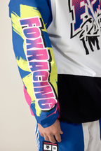 A close-up of a person wearing the 686 Men's Fox Racing Shell Anorak by 686, featuring a blue sleeve with yellow and pink graphics, bold FOX RACING text, and black, white, and blue color sections.