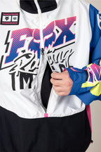A person unzipping a vibrant 686 Men's Fox Racing Shell Anorak, showcasing bold pink, blue, black, and yellow graphics, with their right hand on the zipper.