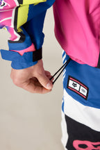 A person pulls the elastic drawstring on a 686 Men's Fox Racing Shell Anorak, featuring vibrant blue, pink, yellow, and white patterns, with a branded patch near their hand.