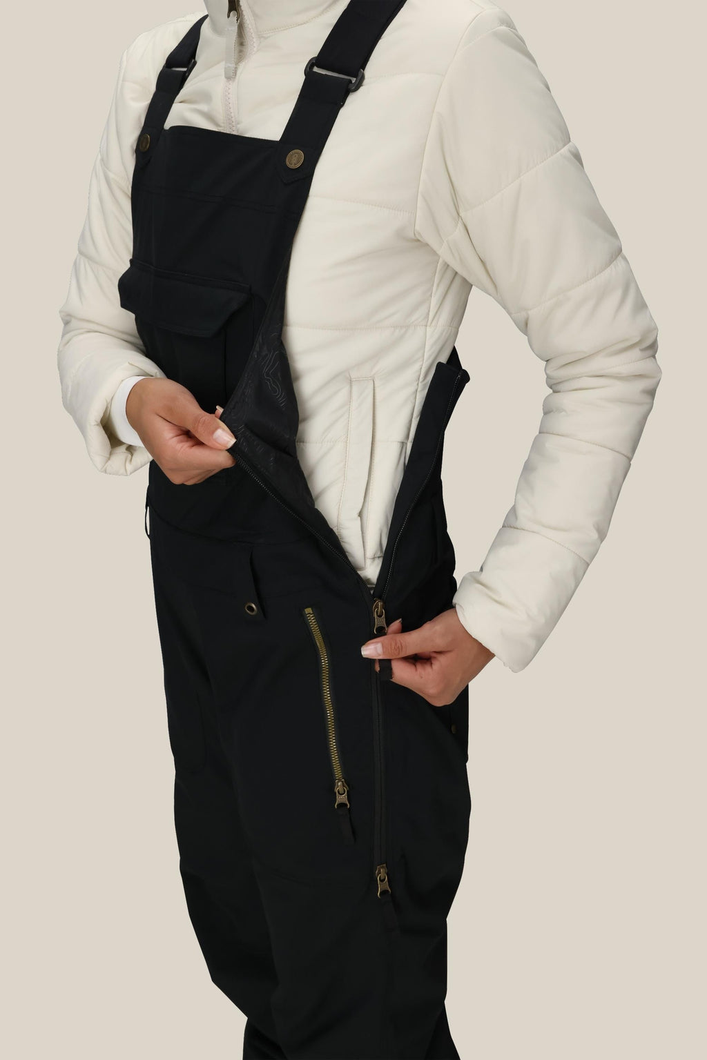 A person wearing a white quilted jacket and 686 Women's Black Magic™ Insulated Bib unzips a side pocket on the bib. The image is cropped from shoulders to knees against a plain background.