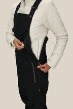 A person wearing a white quilted jacket and 686 Women's Black Magic™ Insulated Bib unzips a side pocket on the bib. The image is cropped from shoulders to knees against a plain background.