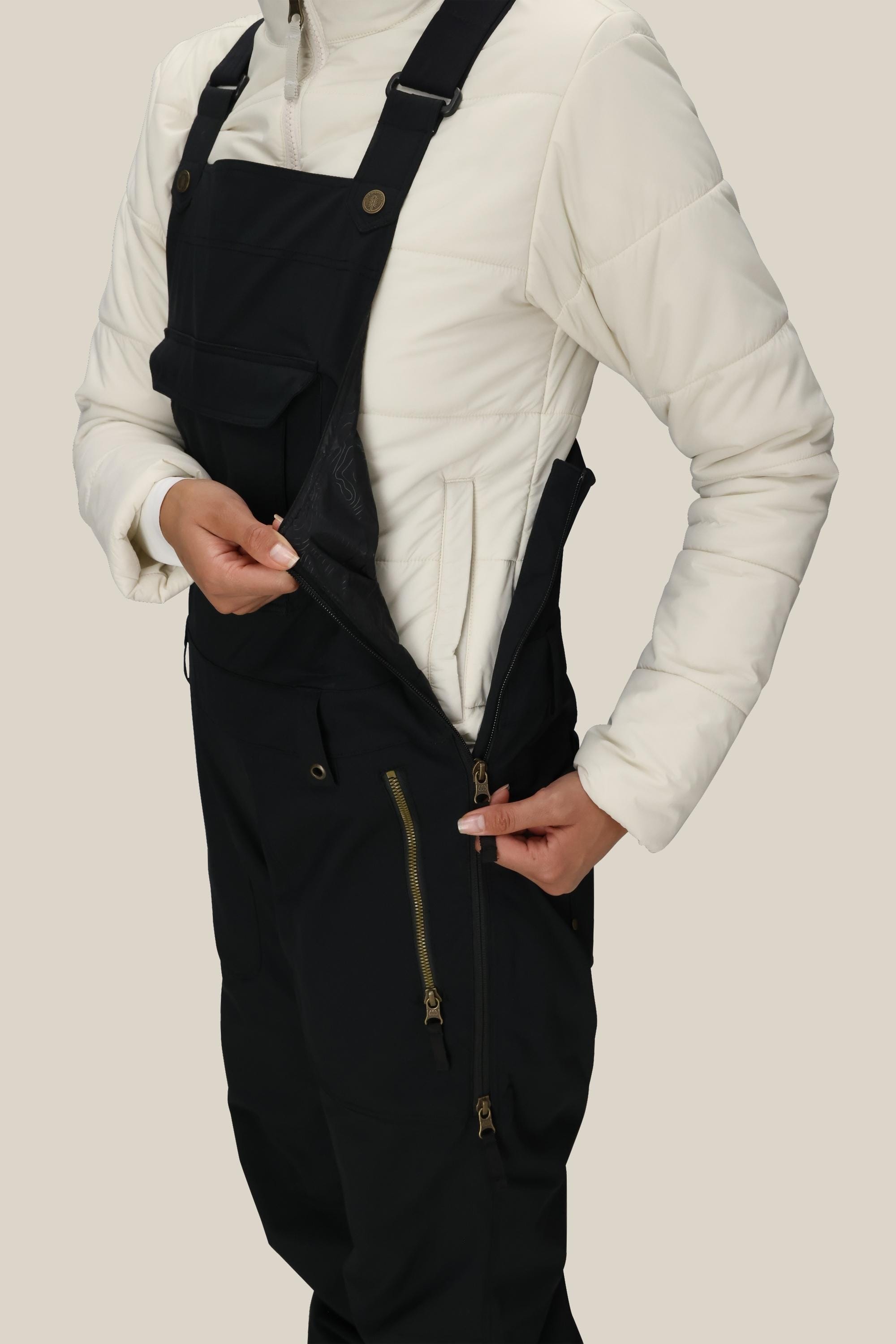 A person wearing a white quilted jacket and 686 Women's Black Magic™ Insulated Bib unzips a side pocket on the bib. The image is cropped from shoulders to knees against a plain background.