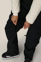 A woman wearing the 686 Women's Black Magic™ Insulated Bib unzips a mesh-lined vent. Black pants and black-and-white sneakers stand out, while a white jacket adds contrast. The photo captures only her lower body and hands.