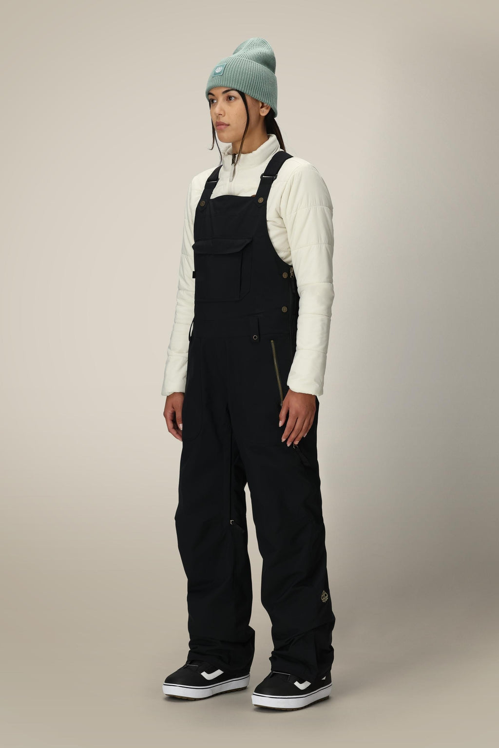 A person stands facing forward, wearing a light blue beanie, a cream quilted top, and 686 Women's Black Magic™ Insulated Bib overalls by 686 with black-and-white sneakers against a plain light background.