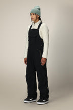 A person stands facing forward, wearing a light blue beanie, a cream quilted top, and 686 Women's Black Magic™ Insulated Bib overalls by 686 with black-and-white sneakers against a plain light background.