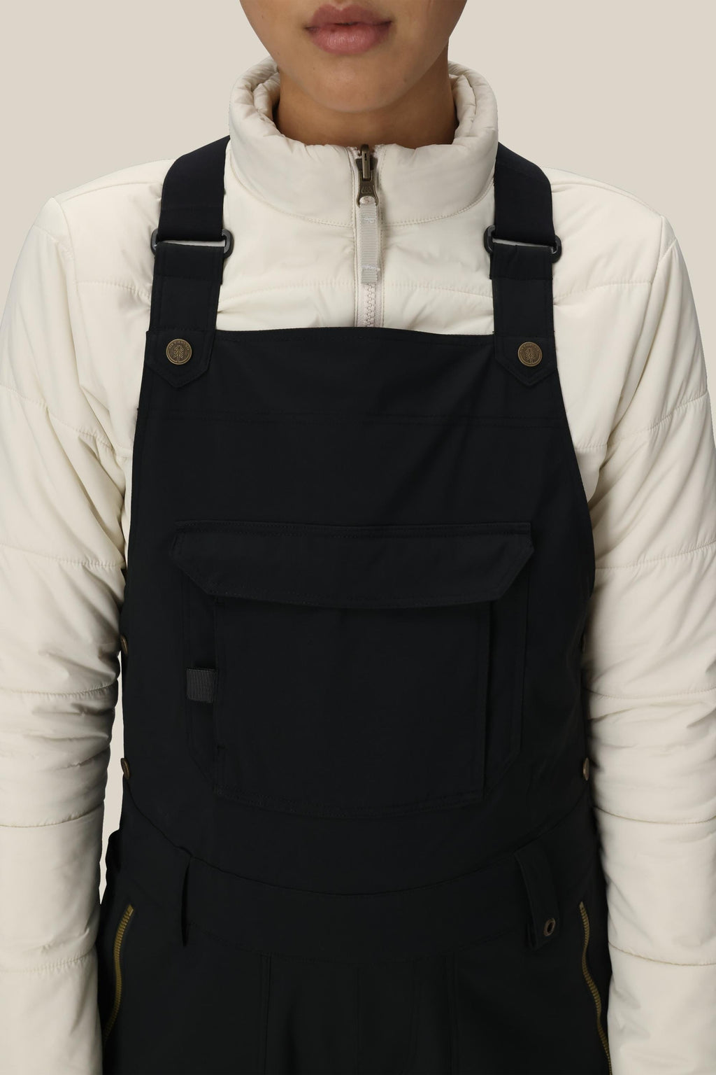 A person is wearing a white zip-up jacket and the 686 Women's Black Magic™ Insulated Bib by 686, shown from shoulders to waist against a plain beige background.