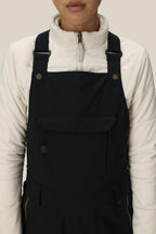 A person is wearing a white zip-up jacket and the 686 Women's Black Magic™ Insulated Bib by 686, shown from shoulders to waist against a plain beige background.