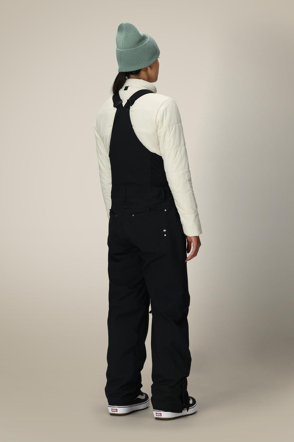 A person stands against a plain beige background, facing away from the camera, wearing a light green beanie, white puffer jacket, and 686 Women's Black Magic™ Insulated Bib by 686 with black and white sneakers.