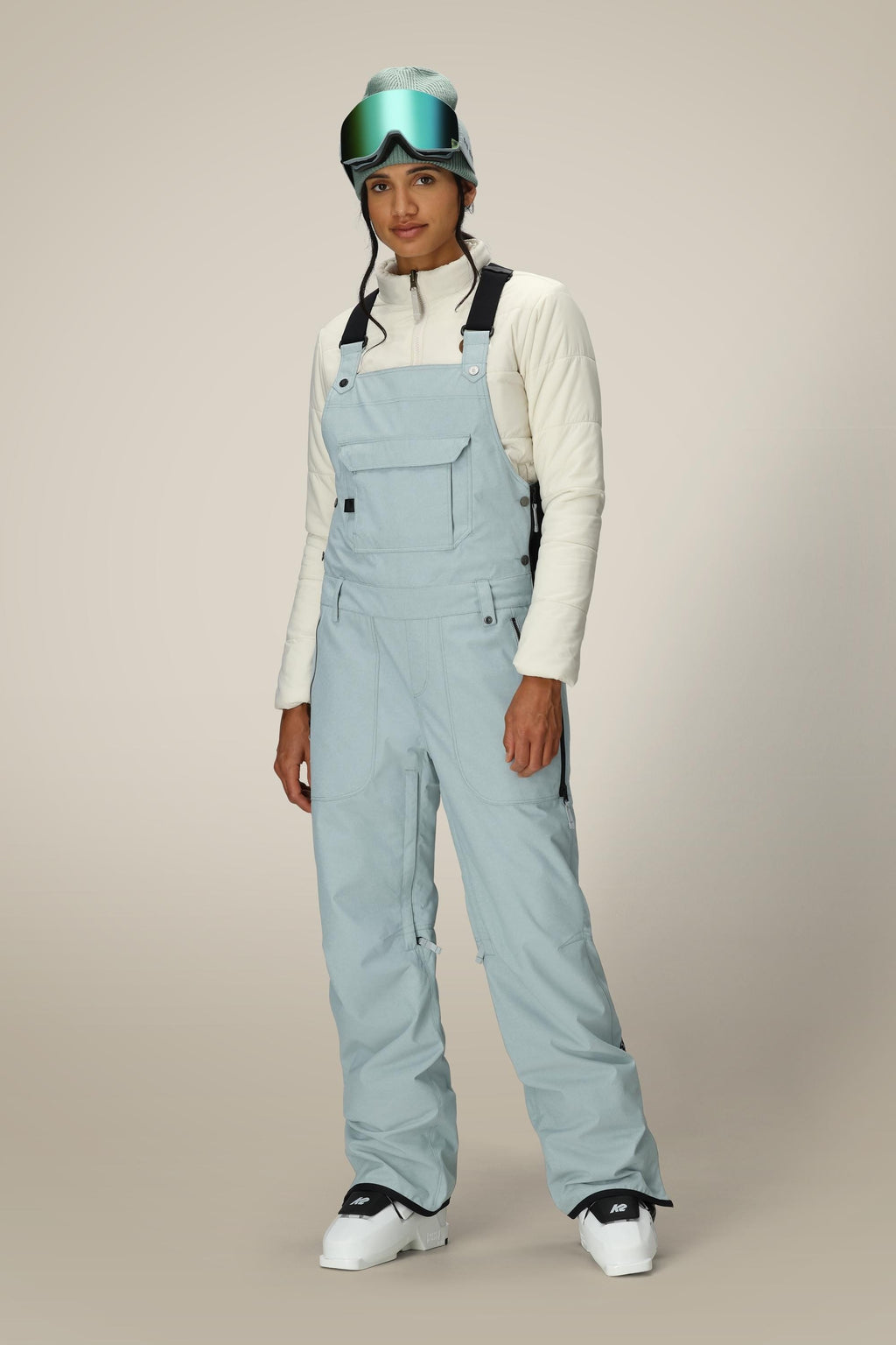 A person wearing the 686 Women's Black Magic™ Insulated Bib stands facing forward in ski goggles, a white jacket, and helmet against a plain beige background.