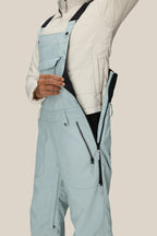 A person wearing the 686 Women's Black Magic™ Insulated Bib in light blue unzips the side to reveal a black inner lining; the image highlights the torso and upper legs against a plain background.