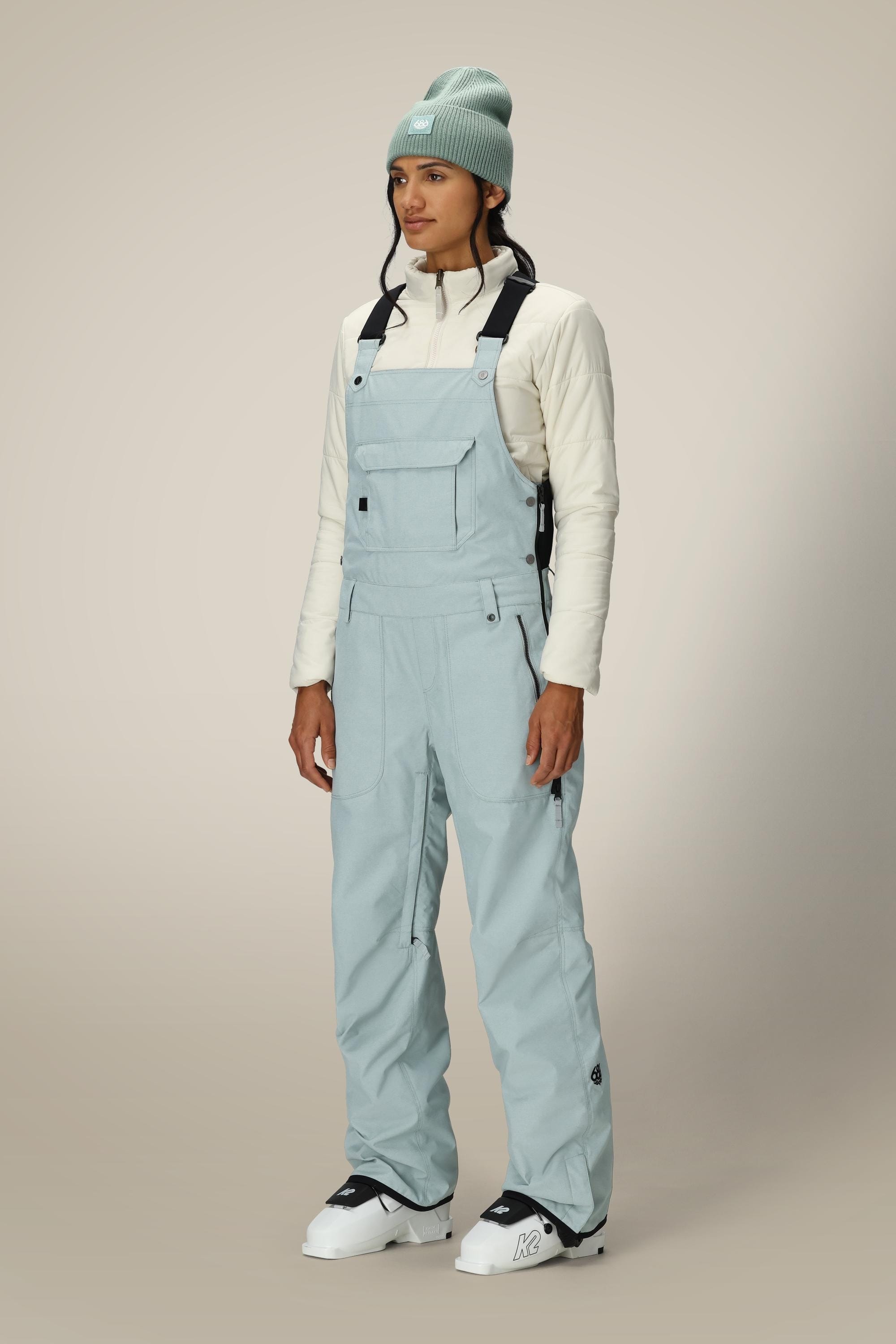 A person stands against a beige background wearing a mint green beanie, cream zip-up jacket, white ski boots, and light blue 686 Women's Black Magic™ Insulated Bib by 686 with black straps.