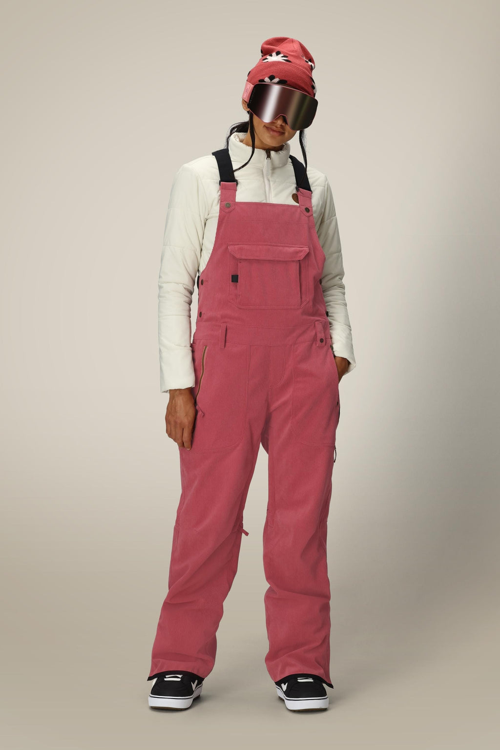 A person stands against a neutral background wearing the 686 Women's Black Magic™ Insulated Bib by 686, paired with a cream jacket, red beanie, large ski goggles, and black-and-white sneakers.