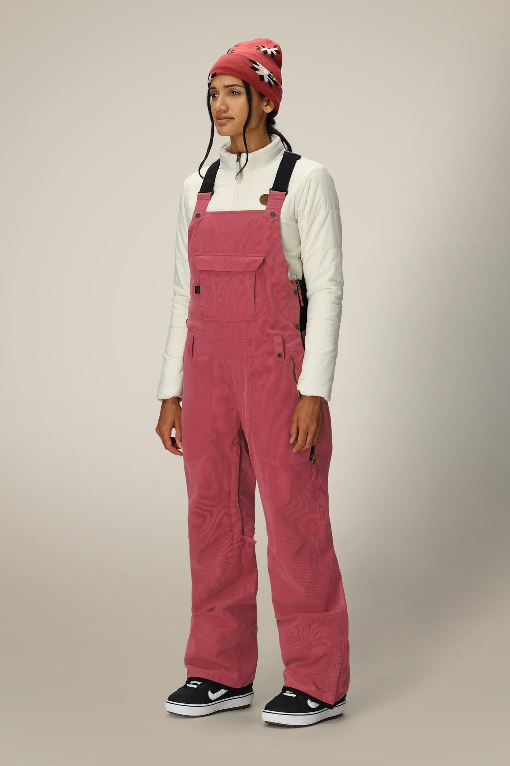 A person stands wearing a white long-sleeve top, the 686 Women's Black Magic™ Insulated Bib by 686, black-and-white sneakers, and a red beanie with a white and black pattern, against a plain light background.