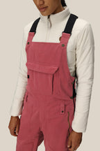 A person wears the 686 Women's Black Magic™ Insulated Bib overalls by 686, featuring pink polyester spandex with black straps and a white quilted jacket, standing against a plain light background. The image is cropped to show torso and upper legs.