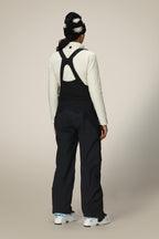 A person stands facing away in a white long-sleeve top, 686 Women's GORE-TEX Dispatch™ Shell Bib overalls by 686, a patterned black beanie, and white ski boots against a plain light background.