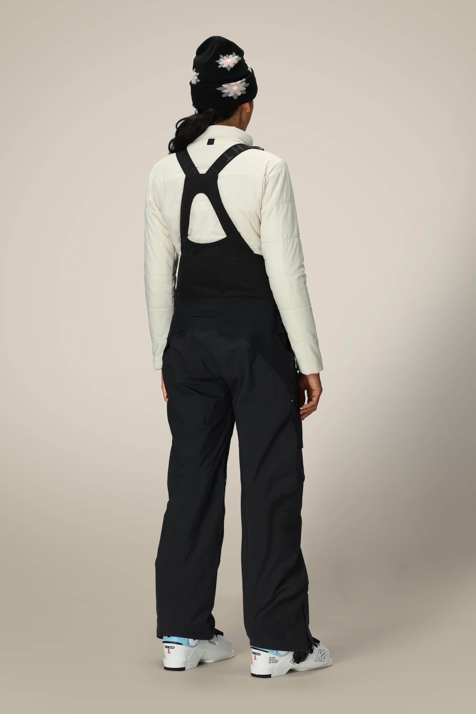 A person stands facing away in a white long-sleeve top, 686 Women's GORE-TEX Dispatch™ Shell Bib overalls by 686, a patterned black beanie, and white ski boots against a plain light background.