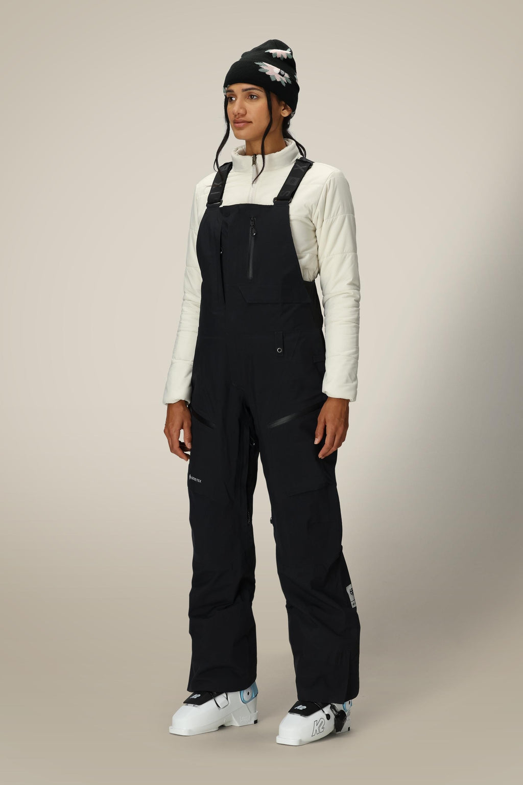 A person wearing 686 Women's GORE-TEX Dispatch™ Shell Bib ski pants, a white quilted jacket, a black beanie with a white pattern, and white ski boots stands against a plain beige background.