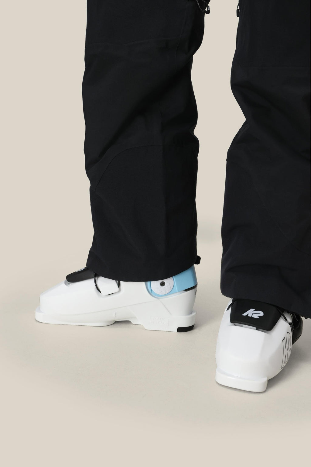 A person wearing 686 Women's GORE-TEX Dispatch™ Shell Bib pants and white ski boots with blue accents, featuring the K2 logo on the heel, stands on a light-colored surface.