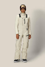 A person wearing the 686 Women's GORE-TEX Dispatch™ Shell Bib with black straps, a front zipper, and multiple pockets, paired with black snow goggles and shoes, stands against a plain light background.