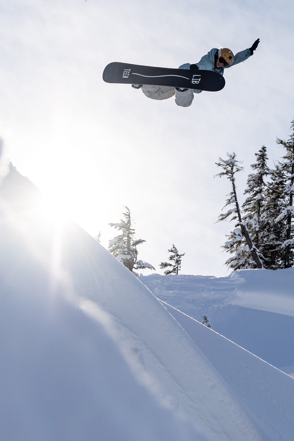 A snowboarder wearing the 686 Women's GORE-TEX Dispatch™ Shell Bib and a helmet catches air above a snowy slope, with sunlight beaming and evergreens in the background.