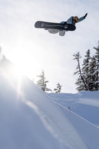 A snowboarder wearing the 686 Women's GORE-TEX Dispatch™ Shell Bib and a helmet catches air above a snowy slope, with sunlight beaming and evergreens in the background.