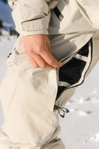 Standing in the snow, a person wearing the 686 Women's GORE-TEX Dispatch™ Shell Bib unzips a mesh vent on the side of their light-colored pants.