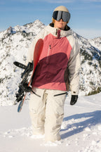 A person wearing the 686 Women's GORE-TEX Dispatch™ Shell Bib stands in deep snow holding a snowboard, with snowy mountains and a clear blue sky in the background.