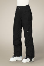 A person wears black 686 Women's GORE-TEX Willow™ Insulated Pants with zippered pockets and reinforced knees, plus white ski boots, standing against a beige background.