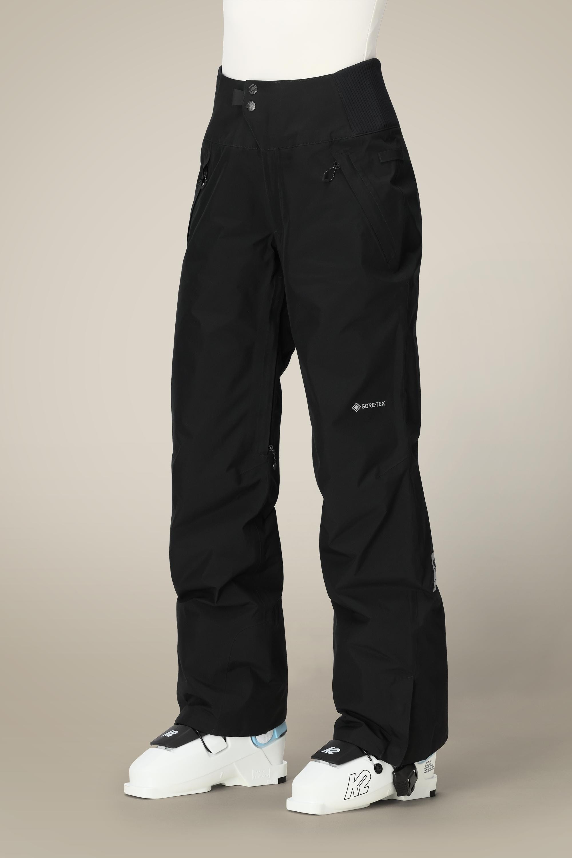A person wears black 686 Women's GORE-TEX Willow™ Insulated Pants with zippered pockets and reinforced knees, plus white ski boots, standing against a beige background.
