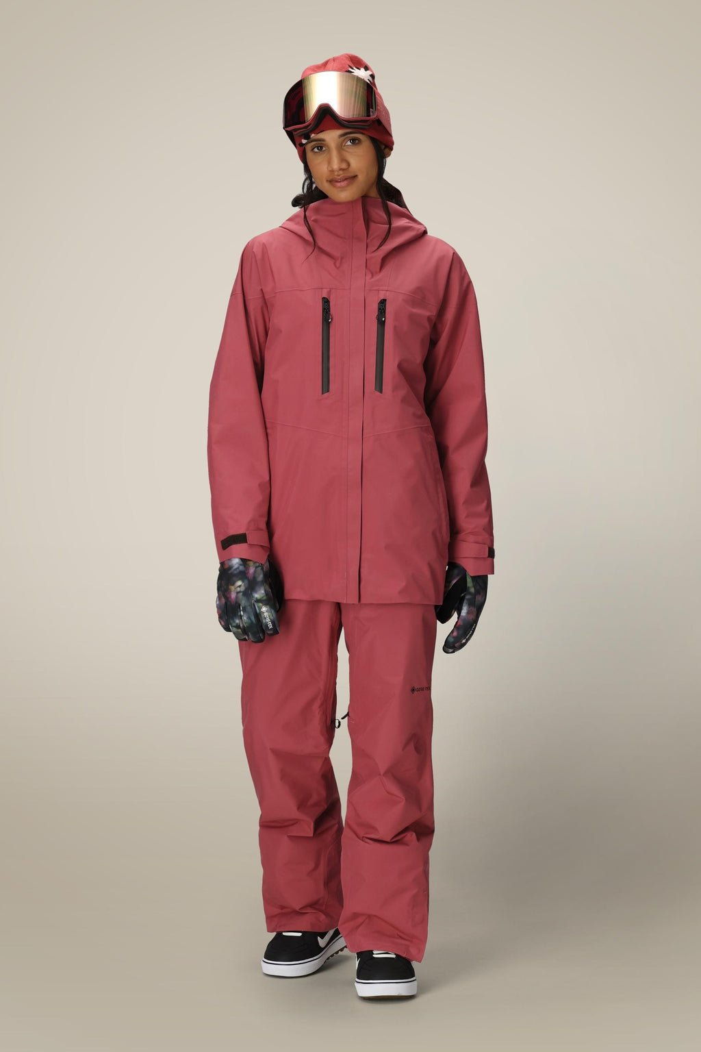 A person stands facing forward, wearing a matching pink ski jacket and 686 Women's GORE-TEX Willow™ Insulated Pant, dark gloves, a red helmet, and ski goggles against a plain light background.