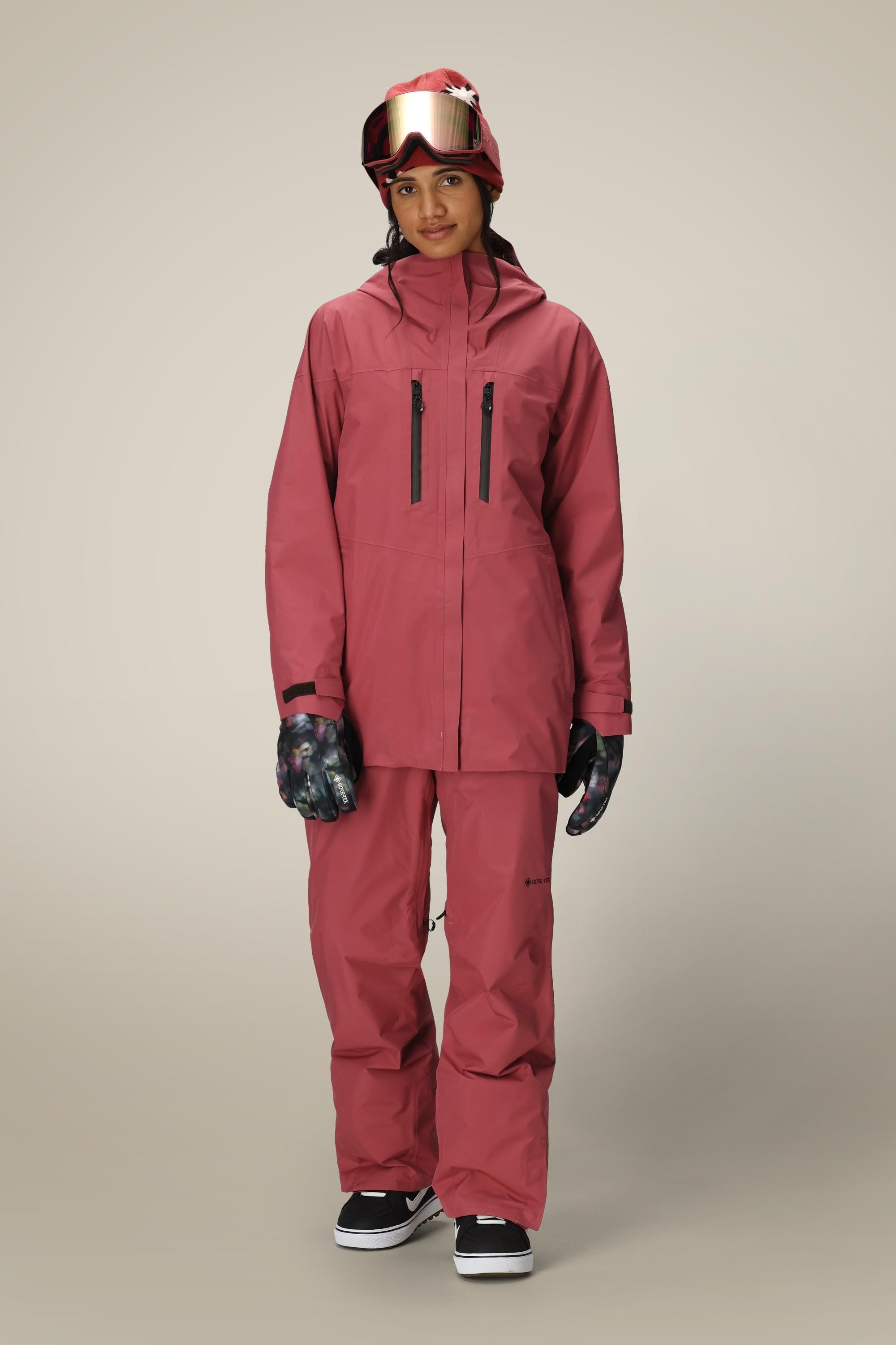 A person stands facing forward, wearing a matching pink ski jacket and 686 Women's GORE-TEX Willow™ Insulated Pant, dark gloves, a red helmet, and ski goggles against a plain light background.