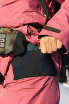 A person in a pink jacket and black ribbed sweater adjusts the 686 Women's GORE-TEX Willow™ Insulated Pant by 686, with a camo-patterned bag strap visible and a wintry, snowy background.
