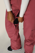 A person wearing the 686 Women's GORE-TEX Willow™ Insulated Pant in pink unzips a side vent, revealing black mesh. Styled with a white long-sleeve shirt and black-and-white sneakers, the look is both functional and fashionable. Brand: 686.