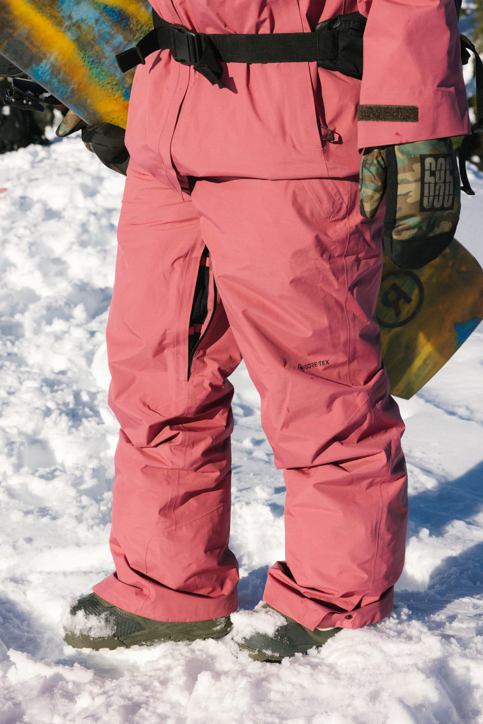 A person wearing 686 Women's GORE-TEX Willow™ Insulated Pants in pink and dark gloves stands on snow, holding a snowboard upright, with only their lower body visible.