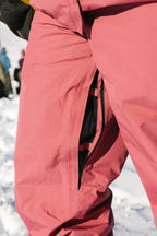 A person wears the 686 Women's GORE-TEX Willow™ Insulated Pant in pink, featuring a partially unzipped side pocket, while standing on snow with blurred figures in the background.