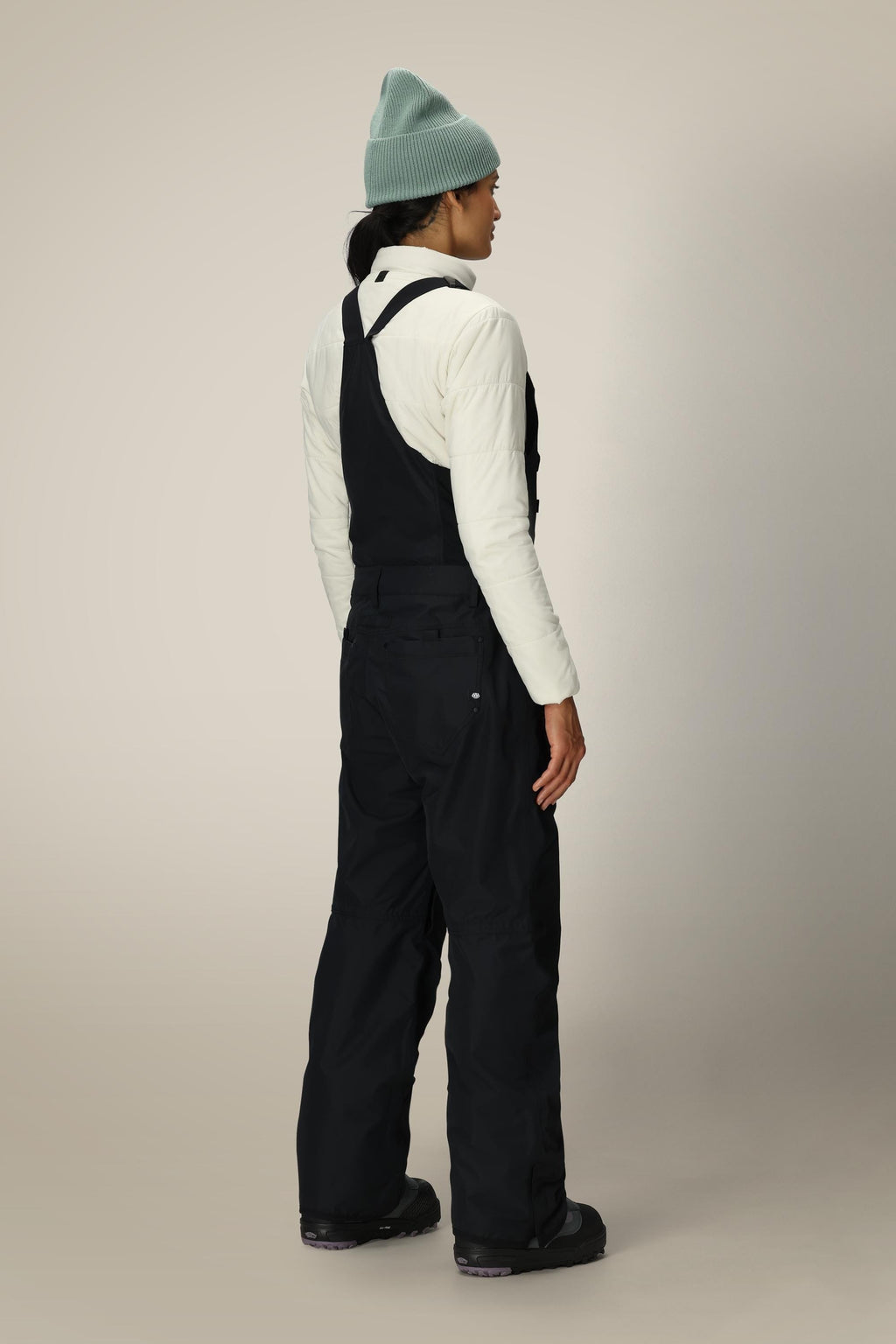 A person stands facing away, wearing a light green beanie, white quilted jacket, black boots, and 686 Black Magic™ 20K Shell Bib women's snow pants by 686, against a plain light background.